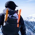 Rear view of a backcountry traveler wearing the Outdoor Vitals Skyline 30 Fastpack packed with winter gear, showing snow safety equipment secured in the side stretch pockets against a snowy mountain background.