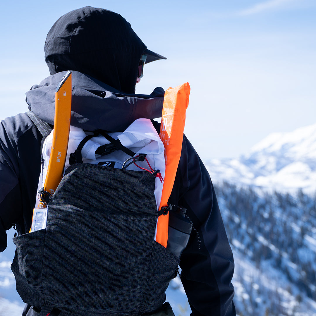Rear view of a backcountry traveler wearing the Outdoor Vitals Skyline 30 Fastpack packed with winter gear, showing snow safety equipment secured in the side stretch pockets against a snowy mountain background.