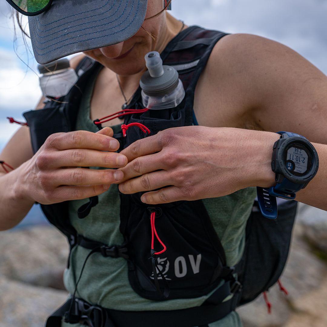 Close-up front view of a trail runner adjusting the sternum straps on the Outdoor Vitals Skyline 30 Fastpack vest harness, showing the dual flask pockets with soft flasks inserted.