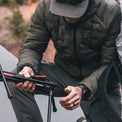 A man wearing a dark gray puffer jacket, gray pants, and a gray cap is sitting outdoors and holding a black and red tripod.