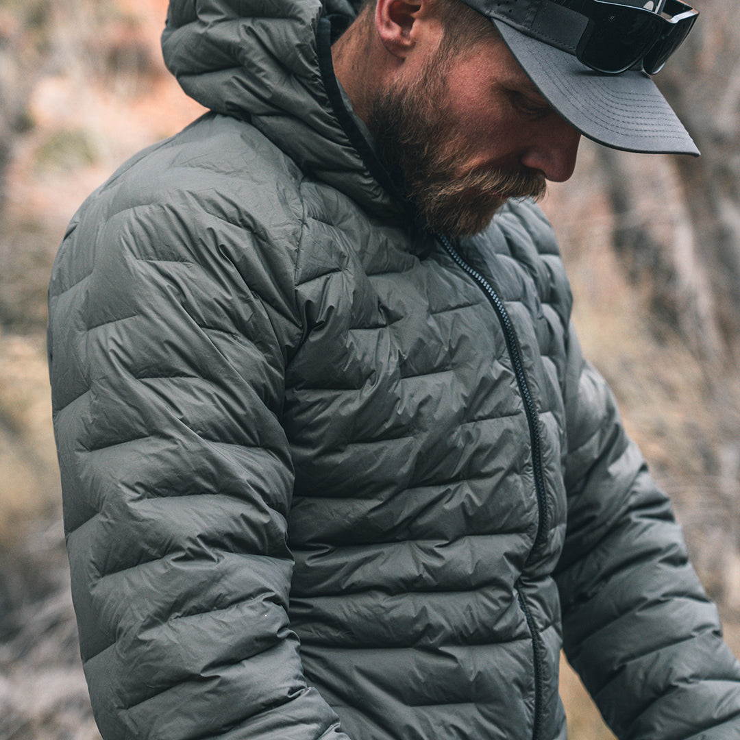 A man wearing a dark gray hooded puffer jacket and a gray baseball cap with sunglasses, photographed outdoors against a blurred natural background.