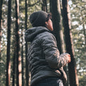 A person standing in a forest wearing a dark gray quilted puffer jacket, a dark gray ribbed knit beanie, and dark pants, holding a small cup.