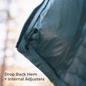 Close-up of a quilted puffer jacket corner with a black zipper, black toggle closure, and text reading 'Drop Back Hem + Internal Adjusters' over a blurred outdoor background.