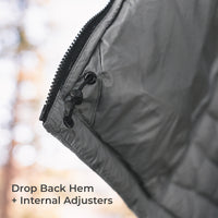 Close-up of a dark gray quilted puffer jacket corner with a black zipper and black plastic adjuster, set against a blurred outdoor background with text reading 'Drop Back Hem + Internal Adjusters'.
