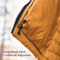 Close-up of a mustard yellow puffer jacket corner with a black zipper, black grommet closure, and text reading 'Drop Back Hem + Internal Adjusters' over a blurred outdoor background.