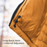 Close-up of a mustard yellow puffer jacket corner with a black zipper, black grommet closure, and text reading 'Drop Back Hem + Internal Adjusters' over a blurred outdoor background.