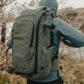 Side-three-quarters view of a hiker wearing a hooded jacket and the Outdoor Vitals KotaUL Ultralight Travel & Adventure Backpack, showing the side water bottle pocket, compression straps, padded hip belt, and Outdoor Vitals branding logo on the front panel.