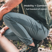 A person outdoors wearing blue athletic jogger-style pants with a small black logo near the lower leg, paired with a dark shirt and hiking shoes. White text at the top reads 'Mobility + Comfort full freedom of motion.