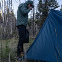 A hiker wearing a hooded jacket and Outdoor Vitals Highline Thermal Leggings standing next to a pitched tent at a backcountry campsite.