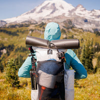 Rear view of a hiker wearing the Outdoor Vitals CS40 Ultra Backpack rigged with a rolled foam sleeping pad, tripod, and gear mesh pocket, facing a snow-capped mountain peak.