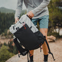 A hiker holding the Outdoor Vitals Skyline 30 Fastpack by its top loop, highlighting the front stretch mesh pocket, shoulder harness water bottle sleeve, and roll-top closure.