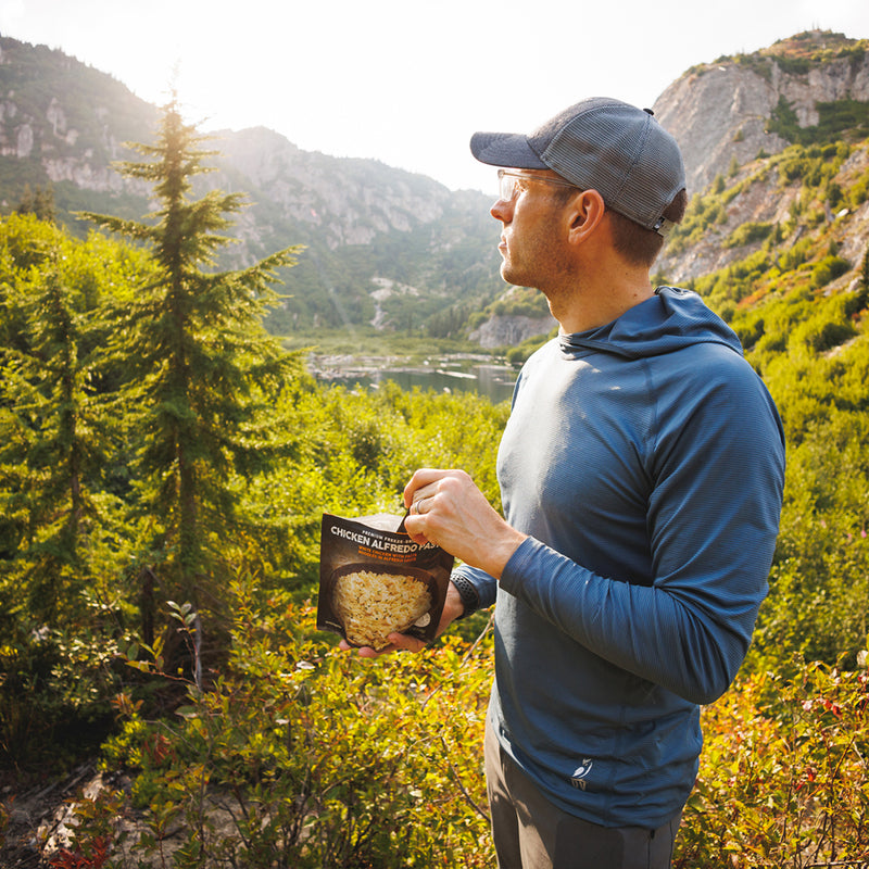 Hiker wearing the Altitude Sun Hoodie enjoying a meal outdoors with mountains and trees in the background.