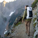 Side profile of a hiker carrying a backpack along a mountain ridge trail, wearing Outdoor Vitals Satu Adventure Pants and a jacket overlooking a scenic valley.