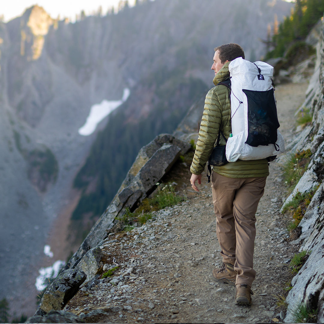 Side profile of a hiker carrying a backpack along a mountain ridge trail, wearing Outdoor Vitals Satu Adventure Pants and a jacket overlooking a scenic valley.