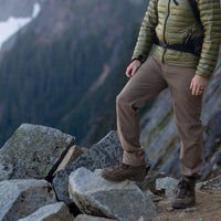Close-up of a hiker standing on rocky terrain wearing Outdoor Vitals Satu Adventure Pants, hiking boots, and a puffer jacket with a mountain backdrop.