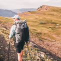 Back view of a hiker in shorts wearing the Outdoor Vitals Shadowlight backpack with a sleeping pad attached, ascending a sunlit alpine ridge.