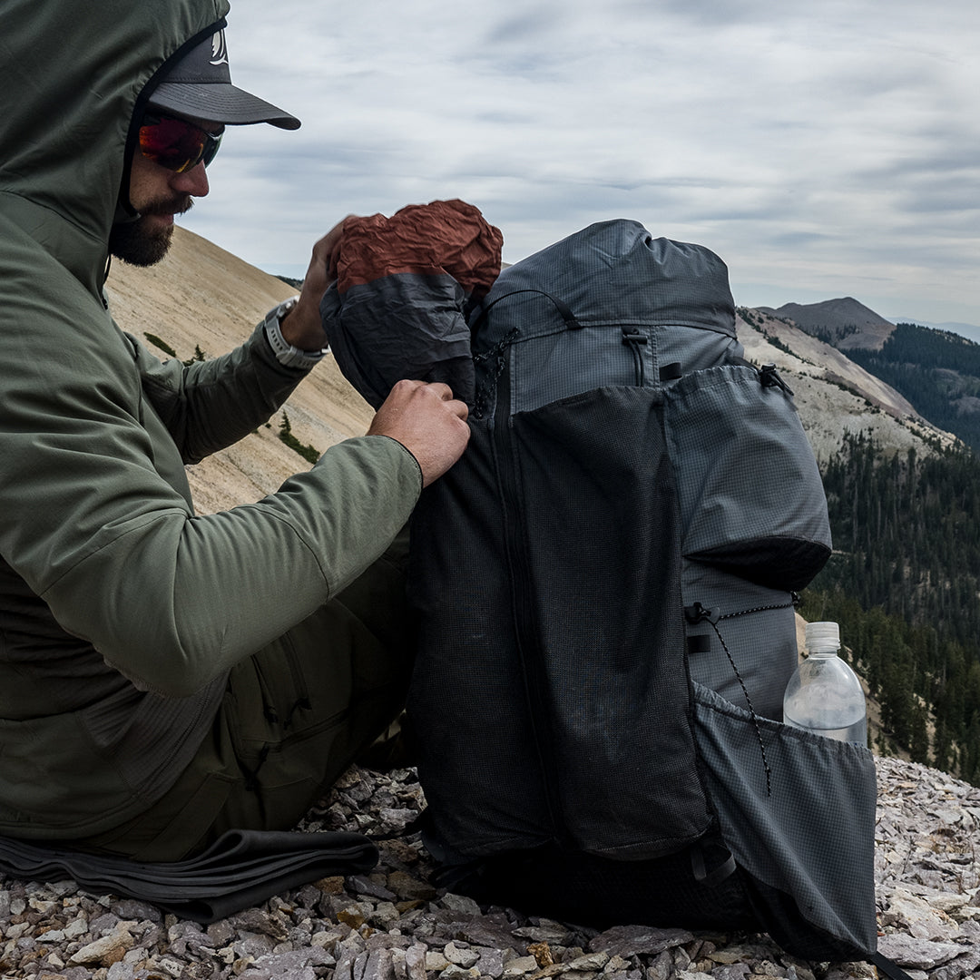 Backpacker in a jacket loading gear into the front mesh pocket of an Outdoor Vitals Shadowlight backpack while sitting on rocky terrain with forested mountains in the background.