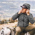 A man wearing a gray puffer jacket with a fitted hood sits outdoors on a rocky mountain slope, adjusting the hood. White text overlays read 'Seals In Heat', 'fitted hood', and 'adjustable hem', with a backpack visible at the lower left.