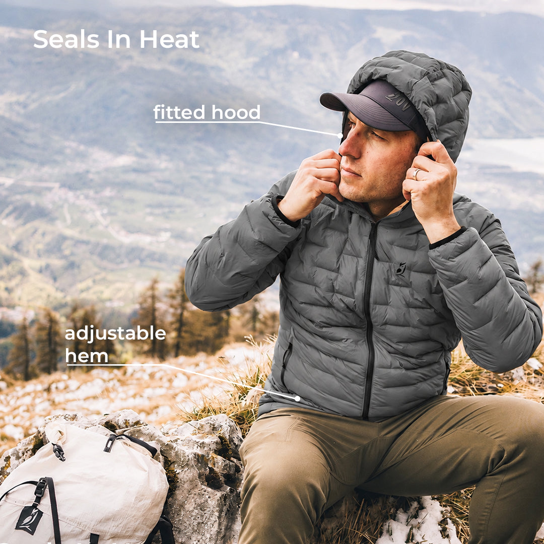 A man wearing a gray puffer jacket with a fitted hood sits outdoors on a rocky mountain slope, adjusting the hood. White text overlays read 'Seals In Heat', 'fitted hood', and 'adjustable hem', with a backpack visible at the lower left.