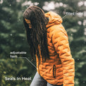 A woman outdoors in falling snow wearing an orange quilted puffer jacket with a fitted hood, with text callouts reading 'fitted hood', 'adjustable hem', and 'Seals In Heat'.