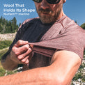 A man outdoors wearing a merino short-sleeve knit shirt and pulling the sleeve outward to demonstrate the fabric's elasticity and shape retention. Text in the top left reads 'Wool That Holds Its Shape NuYarn merino'.