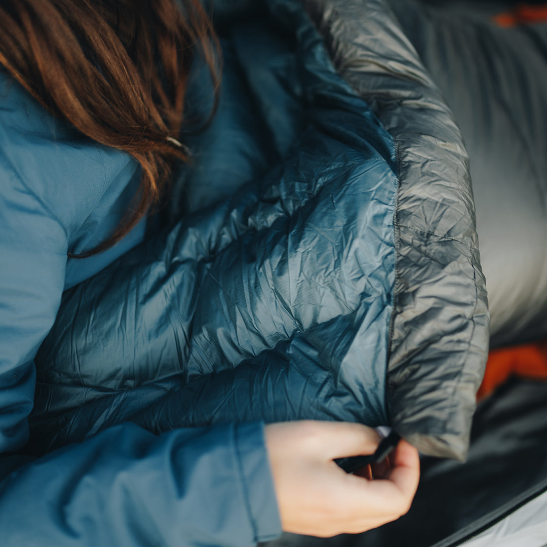 Close-up of a person in a jacket holding the edge of a puffy down top quilt, showing the insulated neck baffle and stitching details.