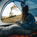 Camper sitting inside a tent in a down top quilt and jacket, holding a cup and looking out at a grassy hillside with trees during sunrise or sunset.
