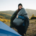 Smiling backpacker in a jacket and beanie draping a top quilt or sleeping bag over their shoulders while standing outdoors in a mountain meadow at sunrise.