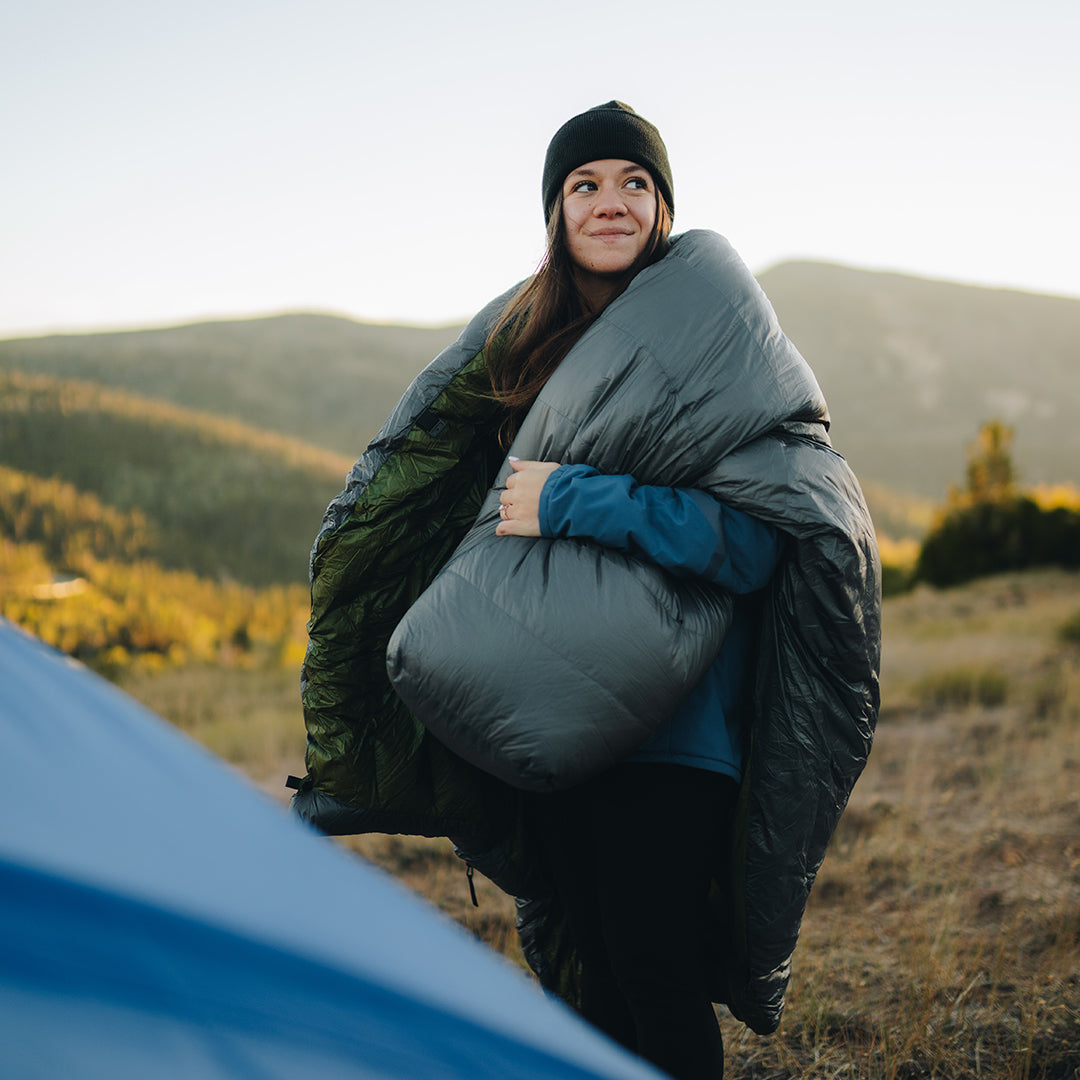 Smiling backpacker in a jacket and beanie draping a top quilt or sleeping bag over their shoulders while standing outdoors in a mountain meadow at sunrise.