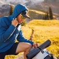 Close-up profile of a male hiker pulling up the hood of his Ventus Active Hoodie while adjusting a rolled sleeping mat strapped to his backpack on a sunny mountain trail.