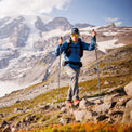 Man hiking uphill in a hooded jacket with a snowy mountain peak in the background.