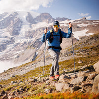 Man hiking uphill in a hooded jacket with a snowy mountain peak in the background.