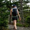 Back view of a female hiker walking through a dense pine forest wearing the Outdoor Vitals Skyline 30 Fastpack, showcasing the large stretch-mesh pocket and roll-top design in an outdoor setting.