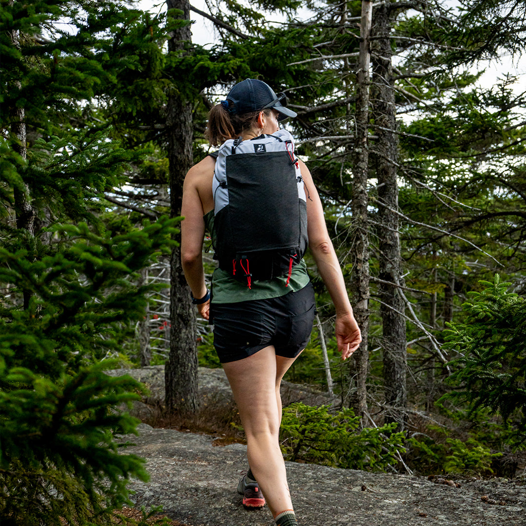 Back view of a female hiker walking through a dense pine forest wearing the Outdoor Vitals Skyline 30 Fastpack, showcasing the large stretch-mesh pocket and roll-top design in an outdoor setting.