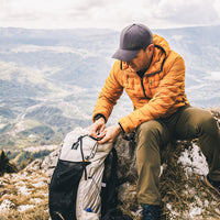 A man in an orange puffer jacket and dark cap sits on a rocky mountain ridge, organizing gear in a light gray and black hiking backpack. The background shows a vast, hazy mountain valley under a cloudy sky.
