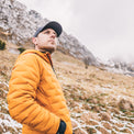 A man wearing a mustard yellow quilted puffer jacket and a black baseball cap stands in a snowy mountain landscape.