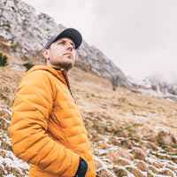 A man wearing a mustard yellow quilted puffer jacket and a black baseball cap stands in a snowy mountain landscape.