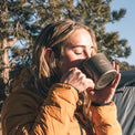 A woman outdoors drinking from a dark gray metal camping mug, wearing a mustard yellow puffer jacket with trees and a tent in the background.