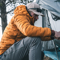 A man wearing a bright mustard yellow quilted puffer jacket, gray pants, and a beige cap crouches next to a gray tent outdoors.