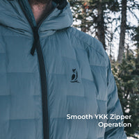 Close-up of a person wearing a puffer jacket outdoors in a snowy forest setting. The jacket is a muted blue-gray color with a black zipper and a small black bird logo on the chest.