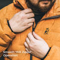 Close-up of a man wearing an orange quilted puffer jacket, adjusting the zipper with both hands. The jacket has a small black bird logo on the left chest and a smooth YKK zipper.