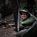 A camper wearing a brimmed hat lounging comfortably inside an Outdoor Vitals Ultralight Complete Hammock System at a wooded campsite.