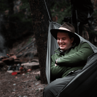 A camper wearing a brimmed hat lounging comfortably inside an Outdoor Vitals Ultralight Complete Hammock System at a wooded campsite.