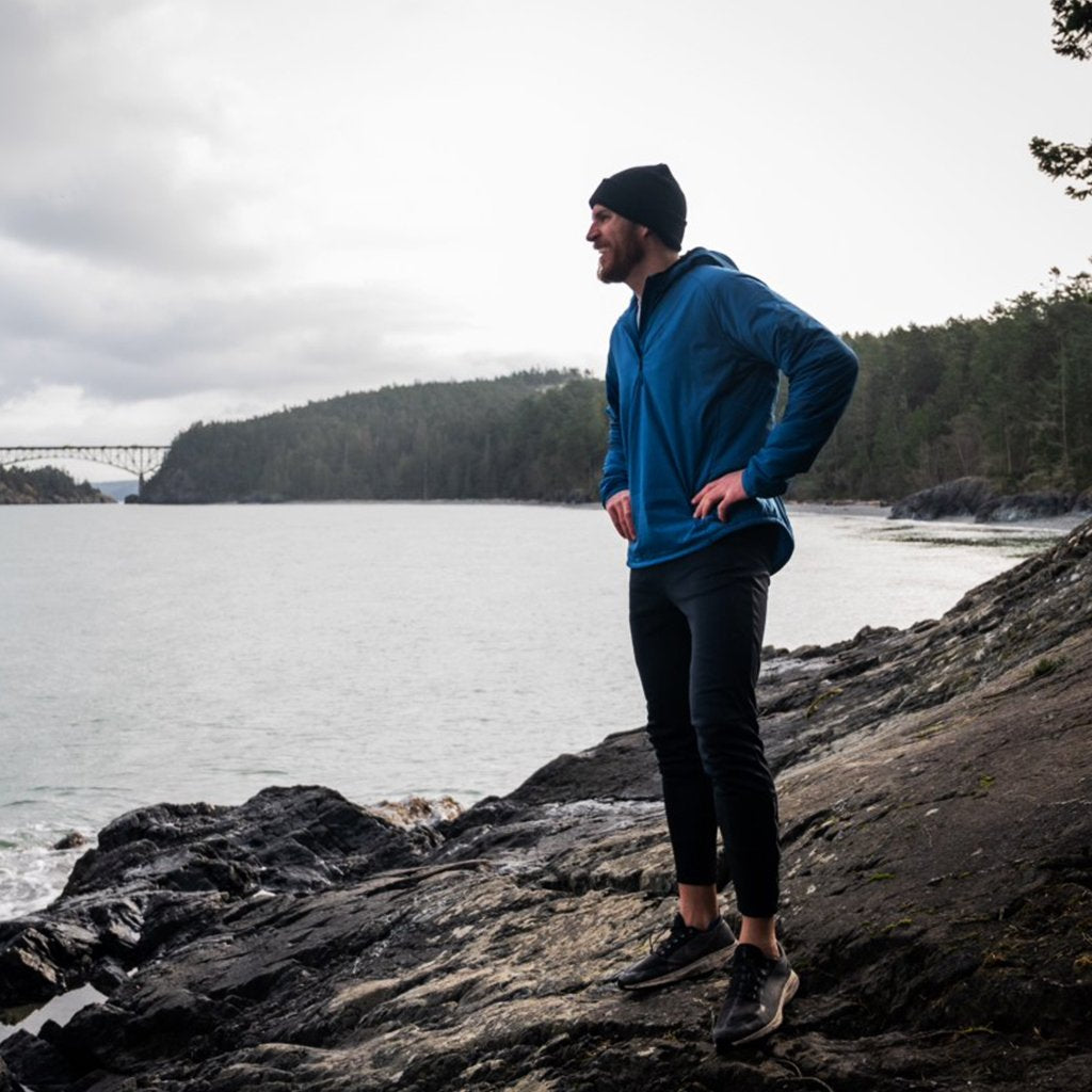 Man standing on a rocky shore wearing a Ventus Active Hoodie and beanie, with a bridge in the distant background.