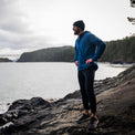 Man standing on a rocky shore wearing a Ventus Active Hoodie and beanie, with a bridge in the distant background.