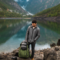 A man wearing a NovaUL down jacket stands by a scenic mountain lake next to a backpacking pack.