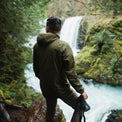 Rear view of a man wearing a Ventus Active Hoodie holding a camera overlooking a forest waterfall.