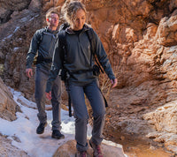 Two hikers wearing Outdoor Vitals Satu Adventure Pants and long-sleeve layers walk along a rocky canyon path with patches of snow.