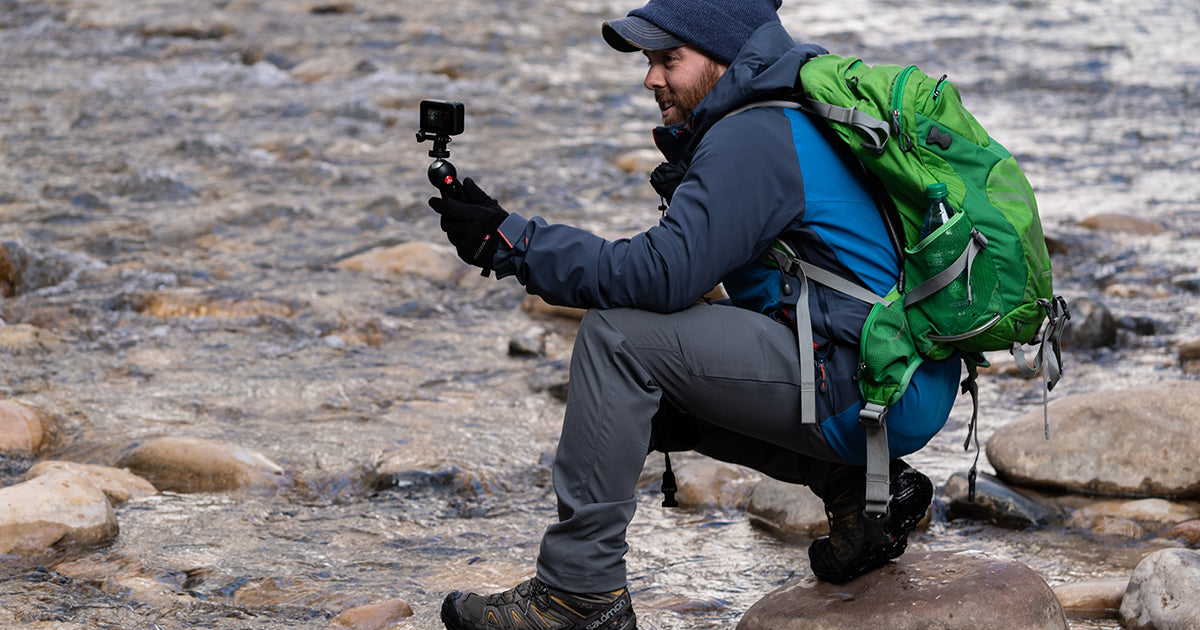 A hiker crouching on rocks near a flowing stream while wearing Outdoor Vitals Satu Adventure Pants and filming with a small action camera on a tripod.