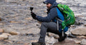 A hiker crouching on rocks near a flowing stream while wearing Outdoor Vitals Satu Adventure Pants and filming with a small action camera on a tripod.